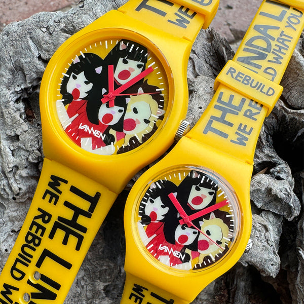 Two yellow The Linda Lindas Vannen Watches with cartoon faces rest on rough wood. This limited edition features straps printed with "THE LINDA WE REBUILD WHAT YOU" and comes in autographed packaging.