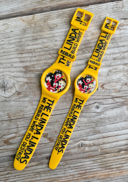 Two yellow The Linda Lindas watches—one adult, one child-sized—with cartoon bands and "WE REBUILD WHAT YOU DESTROY" text rest on wood. Limited edition with precise Japanese quartz movement.