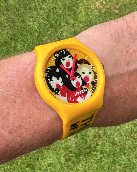 A yellow limited edition The Linda Lindas watch featuring a colorful cartoon bear face on the dial, worn on a wrist against a grassy background.