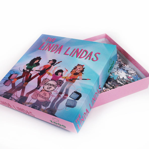 Image of an opened puzzle box against a white background. In a red/pink text with a green outline reads "the linda lindas". Below that is an image of 4 colorful cartoon characters representing the linda lindas playing guitar, bass, and drums. in black text, The bass drum says growing up. The background on the puzzle box is a mix of blue, purple, and green. On the side of the poster box, it says the linda lindas, 1000 piece. The box is pink around the edges.