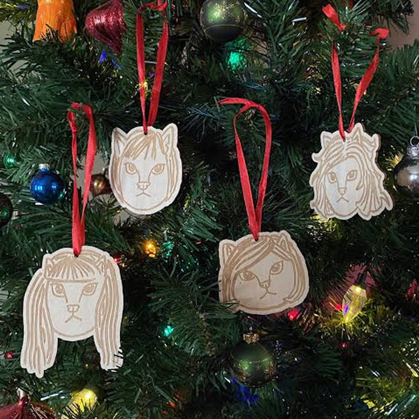 image of four wooden tree ornaments hanging on a christmas tree. each ornament is of the members of the linda lindas as cats