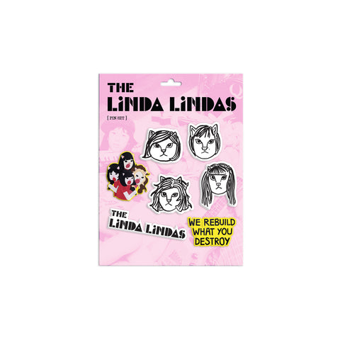 Image of 7 pins against a white background. The cardboard sheet that the pins come attached to is pink. It says the linda lindas in black text. Below that are the pins. 4 of the pins are black and white drawings of cat faces with hair- one cat has longer hair. One pin of the four cats singing- they have black and brown hair and are wearing red shirts. One pin says the linda lindas in black with white outline, and the last pin has a yellow outline with black writing that says "we rebuild what you destroy".